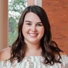 a woman wearing a white off the shoulder top is smiling in front of a brick wall .